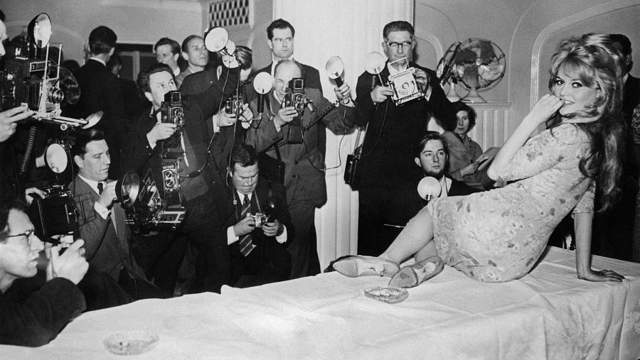 En una sesión fotográfica, una mujer rubia con un vestido estampado posa sobre una mesa, rodeada de fotógrafos con cámaras antiguas. La escena, en blanco y negro, captura un momento de concentración y elegancia.