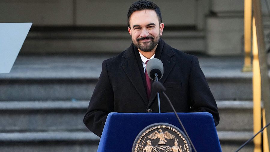Zohran Mamdani, con barba y cabello oscuro, pronuncia un discurso en su investidura. Viste formalmente y se encuentra detrás de un atril con el sello de la ciudad, frente a un micrófono, con el Ayuntamiento de fondo.
