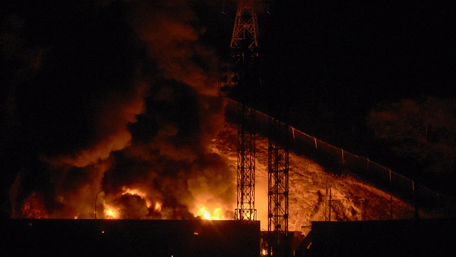 Una escena nocturna de un incendio masivo, con llamas voraces y una gran columna de humo. Se observa una torre de alta tensión en el fondo y estructuras en la parte inferior de la imagen.