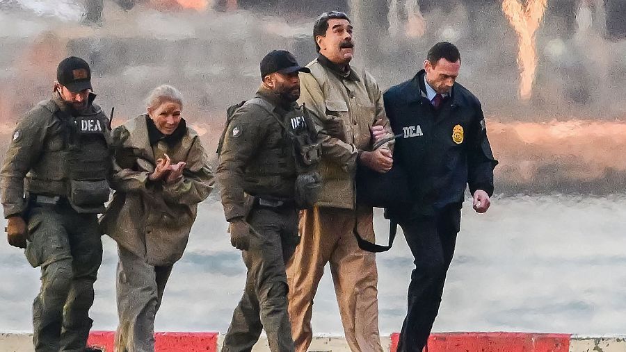 Nicolás Maduro, esposado y vestido con chaqueta beige, es custodiado por agentes de la DEA. Se observa a otros agentes de la DEA, algunos con uniformes tácticos, en un entorno urbano.