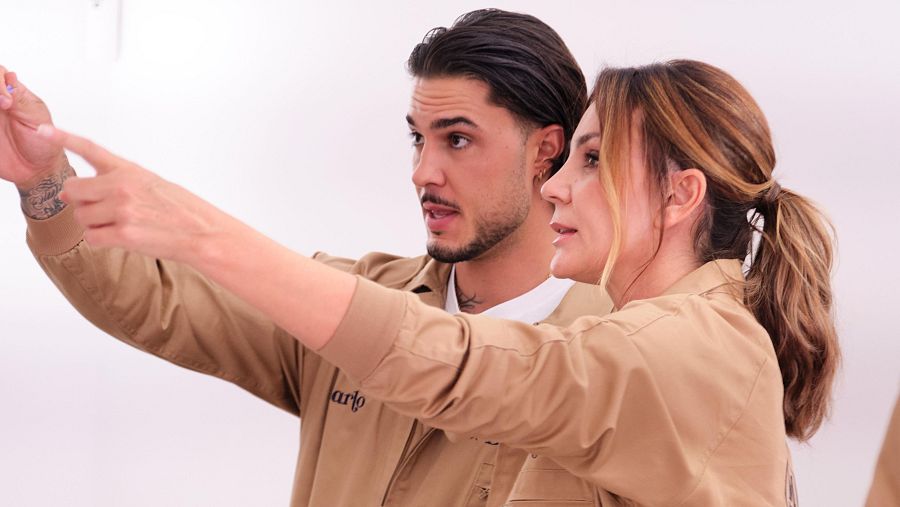 Un hombre con tatuajes y barba, y una mujer con coleta, posan juntos. Ambos visten camisas claras y miran hacia la izquierda, señalando con sus manos.