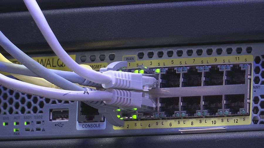 Un panel de red con cables de diferentes colores conectados a puertos etiquetados con números y abreviaturas, con luces LED verdes encendidas y diversas etiquetas como 