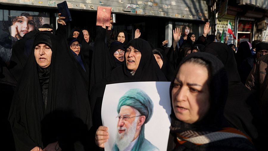 Un grupo de mujeres iraníes, la mayoría con hijab negro, participan en una manifestación en Teherán, algunas mostrando apoyo y sosteniendo objetos, con la bandera de su país al fondo.