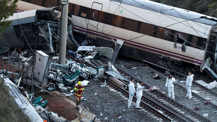 En Adamuz, España, un tren de color blanco con una franja morada de Renfe ha descarrilado, mostrando daños y estando parcialmente volcado. Se observa a bomberos y personal con trajes de protección en la zona del accidente, que se encuentra en un terreno pedregoso.