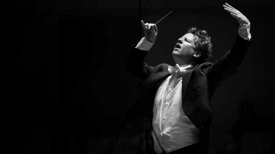 Un director de orquesta, vestido formalmente, dirige con entusiasmo, sosteniendo una batuta y con la mirada alzada, en una imagen en blanco y negro.