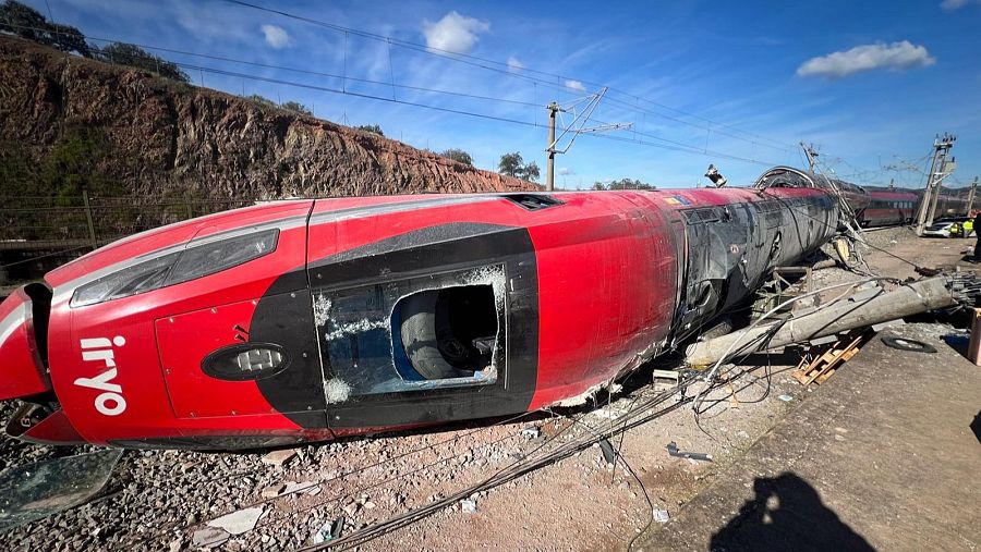 Un tren rojo Iryo descarrilado, con el logo visible, muestra daños significativos yace cerca de una zona con vegetación, con otro tren a lo lejos.