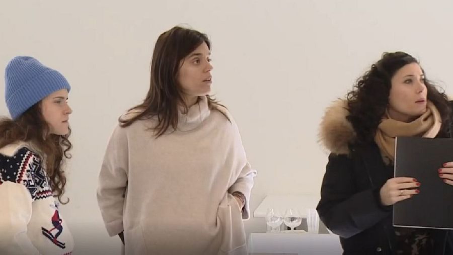 Tres mujeres en un interior con paredes blancas: una con gorro azul y jersey blanco, otra con jersey crema y la tercera con abrigo negro, bufanda beige y carpeta.