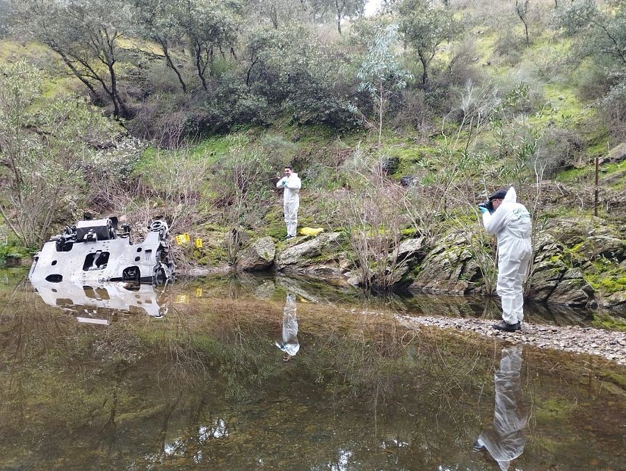 Un río refleja los restos de un tren accidentado, mientras dos personas con trajes blancos, posiblemente agentes, inspeccionan la escena, tomando fotografías y con marcadores amarillos en el entorno natural.