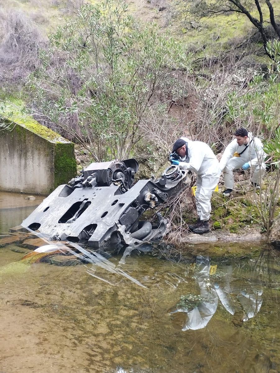 Dos personas con trajes protectores blancos examinan los restos de un tren parcialmente sumergido en un río. La escena muestra fragmentos grises oscuros dañados, con uno de los investigadores inclinado y el otro sentado en la orilla, rodeados de vegetación y una estructura de hormigón.