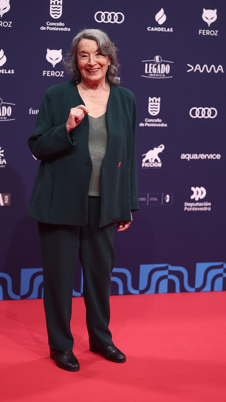 Petra Martínez, con cabello gris, sonríe a la cámara en una alfombra roja, luciendo un traje verde oscuro con una blusa brillante. El fondo muestra varios logos.