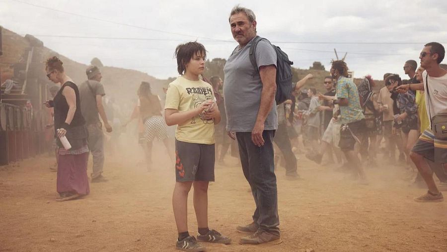 Un hombre y un niño, vestidos con ropa casual, miran hacia el frente. Detrás de ellos, una multitud se mueve en un ambiente polvoriento, con el cielo nublado, sugiriendo un evento al aire libre.