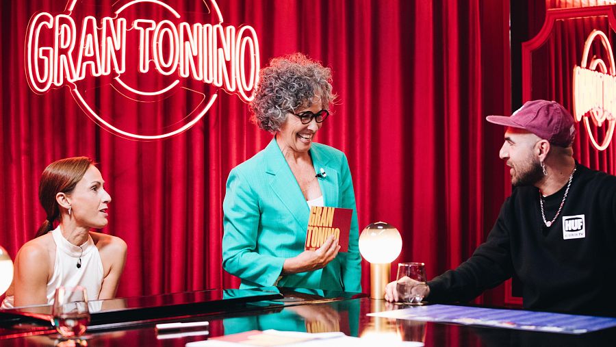 Tres persones comparteixen espai en un estudi amb cortines vermelles i un rètol lluminós. Una dona amb el cabell recollit, una altra amb ulleres i un home amb gorra rosa, tots mirant cap al centre, possiblement durant una conversa o presentació.