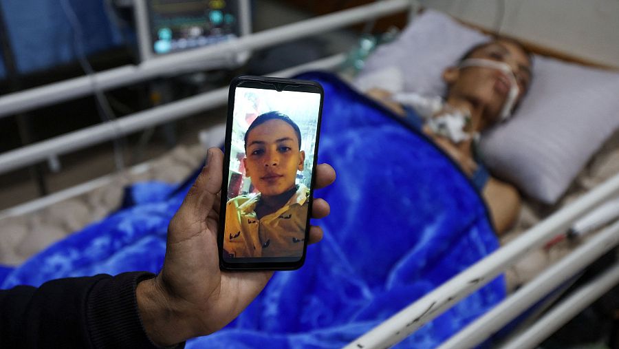 En una habitación de hospital, un paciente conectado a un respirador y otros equipos médicos es fotografiado. En primer plano, un teléfono móvil muestra una imagen del mismo paciente con mejor aspecto.