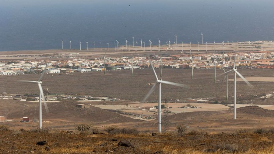 Un paisaje con aerogeneradores en primer plano, ubicados en un terreno seco y con poca vegetación, con una ciudad costera y el mar al fondo.