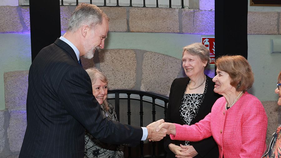 Felipe VI, Rey de España, estrecha la mano de una mujer con chaqueta rosa en un evento. A su lado, otra mujer lleva una chaqueta estampada, mientras que una tercera, detrás de la primera, viste una chaqueta negra sobre un vestido a juego.