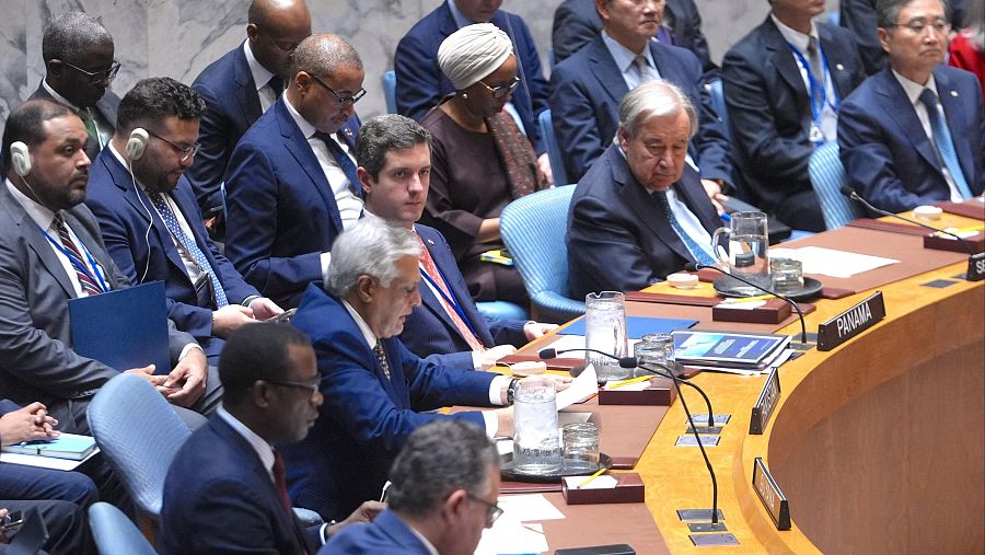 Una sala de reuniones con múltiples personas sentadas alrededor de mesas, incluyendo a un individuo. Se observa la palabra 