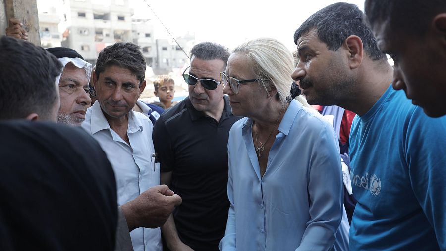 Sigrid Kaag, representante de la ONU, inspecciona una zona y dialoga con residentes. La imagen muestra a Kaag con vestimenta formal, rodeada de hombres, algunos con vestimenta tradicional, en un entorno con edificios dañados.