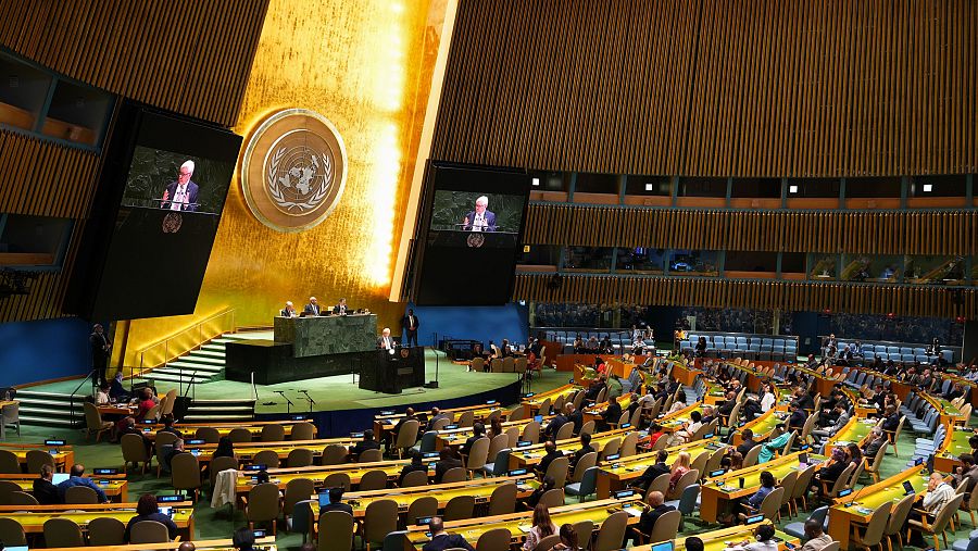 El salón de la Asamblea General de las Naciones Unidas está lleno de delegados. En el estrado, un orador se dirige a la asamblea, con su imagen proyectada en dos grandes pantallas.