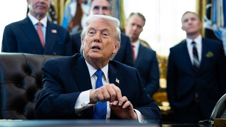 Donald Trump, con traje oscuro y corbata, firma documentos en el Despacho Oval, rodeado de personas vestidas formalmente, con banderas y cortinas doradas como telón de fondo.