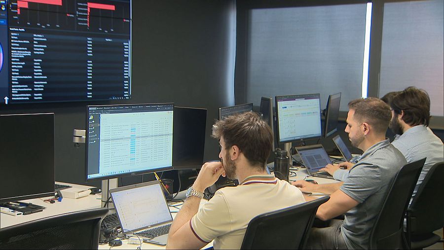 Tres hombres en una sala de control operan con ordenadores, incluyendo portátiles y monitores de escritorio, mientras una pantalla grande muestra datos y gráficos.