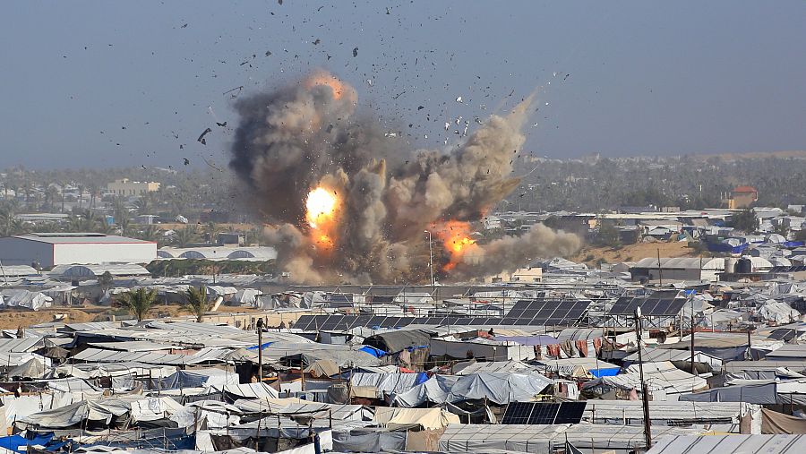 Una gran explosión de color naranja y gris sacude un campamento de refugiados, lanzando escombros al aire. Se observan numerosas tiendas de campaña y estructuras precarias en el campamento, con algunos edificios al fondo.