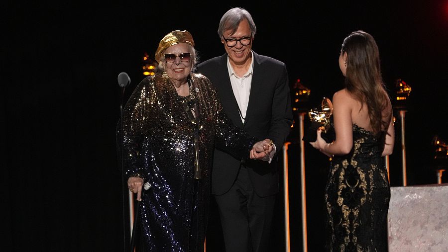 Joni Mitchell, con un vestido brillante y sombrero dorado, recibe un premio, acompañada por un hombre y una mujer en un evento en Los Ángeles.