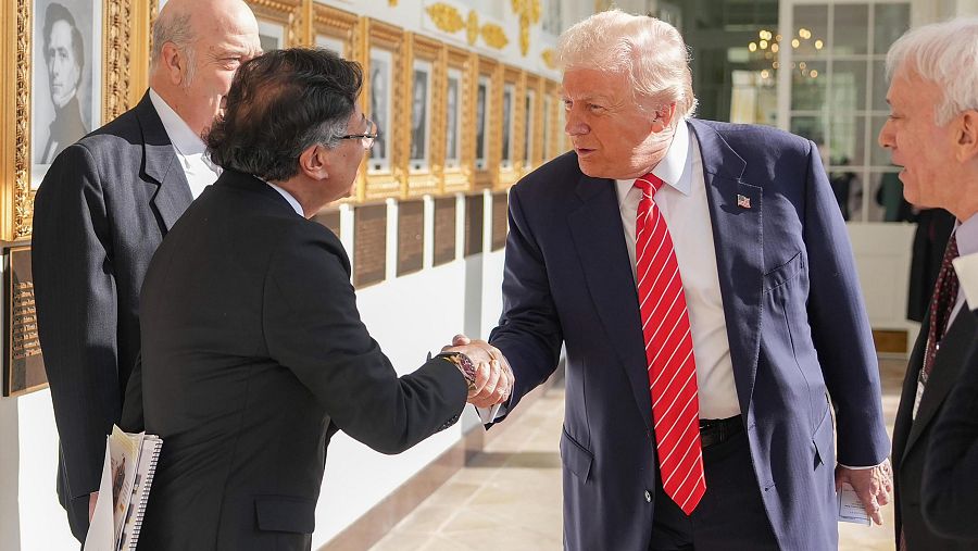 Donald Trump y Gustavo Petro se saludan en la Casa Blanca. Trump lleva un traje azul marino con corbata roja, y Petro un traje negro. Se aprecian otras personas y una pared con cuadros.