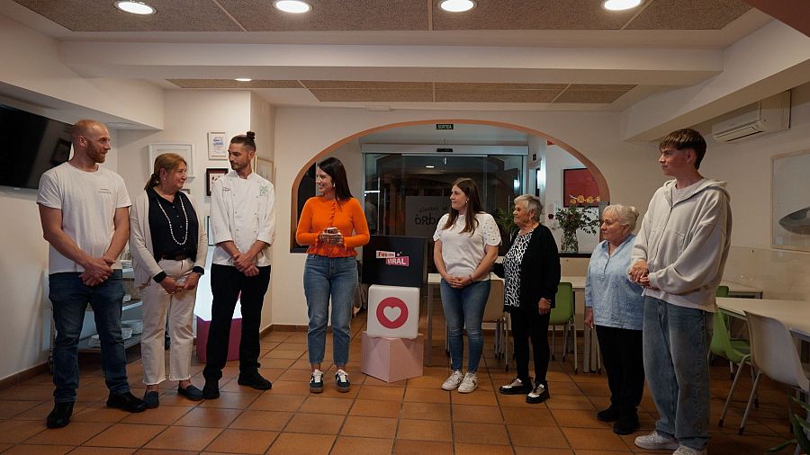 Un grupo diverso de personas, incluyendo un chef y varias mujeres mayores, se reúne en un espacio interior con mesas y una puerta arqueada. Una mujer sostiene un objeto pequeño frente a un cubo con un logo.