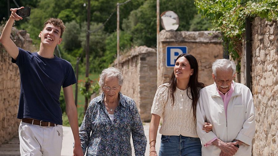 Quatre persones passegen per un carrer empedrat d'un poble. Un jove fa una foto, una dona gran porta ulleres i una brusa florida, una noia vesteix un jersei crema i un home gran una camisa rosa.