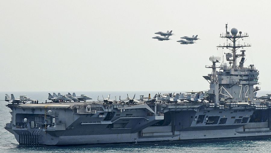 Un portaaviones gris navega por el mar, con aviones de combate en su cubierta y otros cuatro en formación en el cielo despejado.