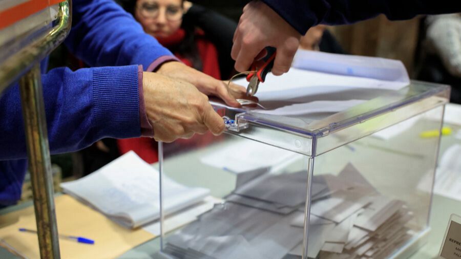 Una urna transparente, repleta de sobres, es abierta con alicates por una persona con camisa azul en el Ayuntamiento de Zaragoza, mientras otra persona observa el proceso.