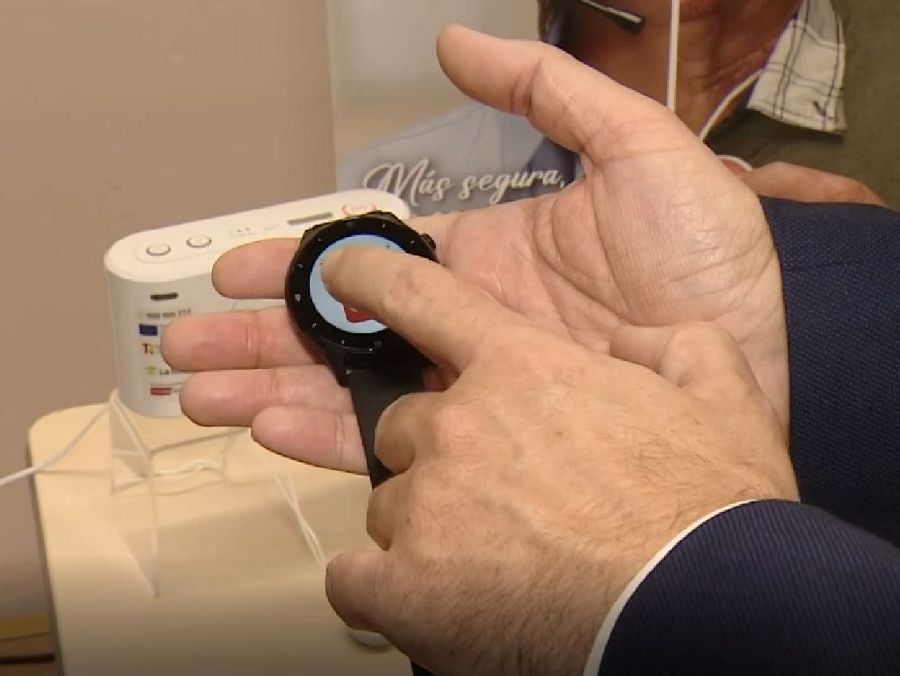 La imagen muestra una mano interactuando con un reloj inteligente de correa negra, cuya pantalla exhibe un icono rojo. En el fondo, se aprecia un dispositivo blanco con indicadores y un cartel con la frase 
