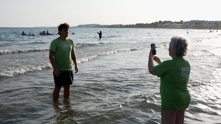 En una platja assolellada, un home jove amb samarreta verda i pantalons curts posa a l'aigua mentre una dona fa fotos. Al fons, el mar i persones gaudint de l'aigua.