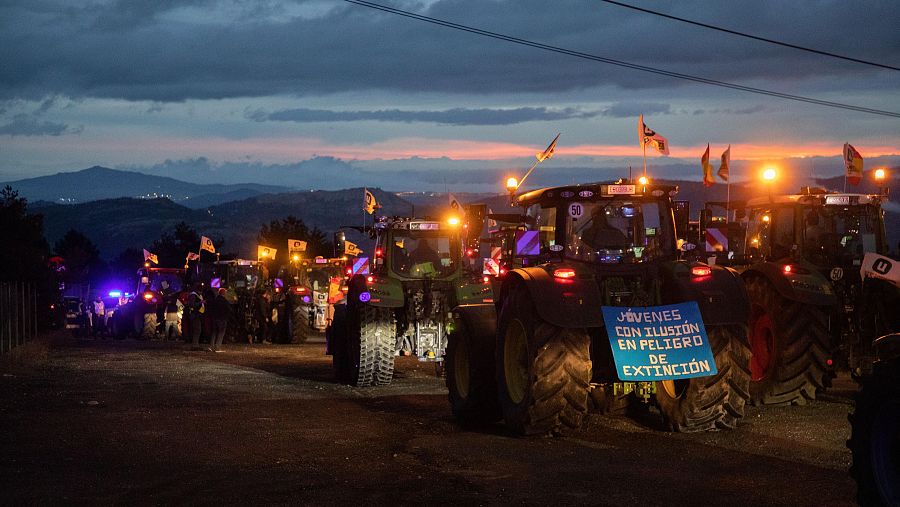 Varios tractores verdes, con las luces encendidas y banderas, se manifiestan al atardecer. Destaca una pancarta azul con un mensaje sobre la juventud. El cielo muestra tonos anaranjados y grises.