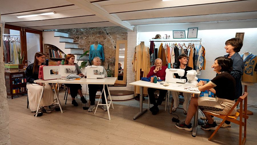 En un taller de costura rústic, un grup de persones de diverses edats treballen amb màquines de cosir, possiblement en una activitat supervisada per una dona amb vestit blau.