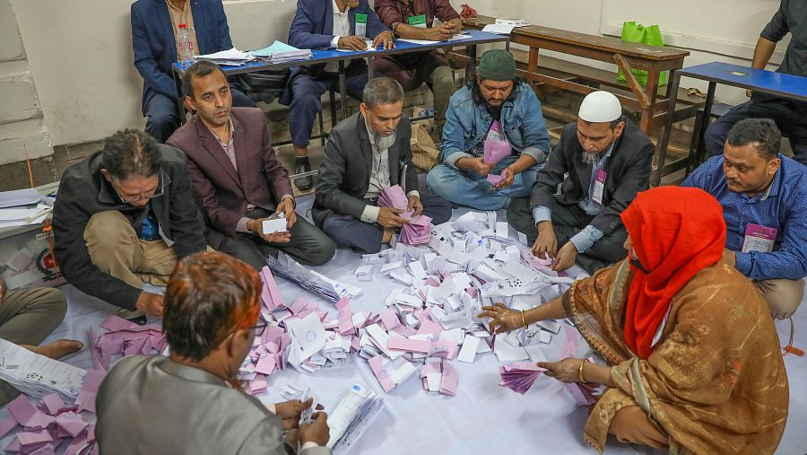Un grupo de personas, incluyendo hombres con trajes y ropa informal, y una mujer con pañuelo rojo, se encuentran contando votos en el suelo, rodeados de papeletas de colores.