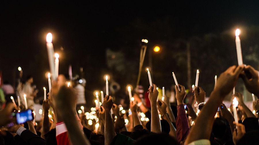 Una multitud se congrega en la noche, iluminada por la luz de las velas. Una bandera ondea en medio de la gente, mientras alguien documenta la escena con un teléfono.