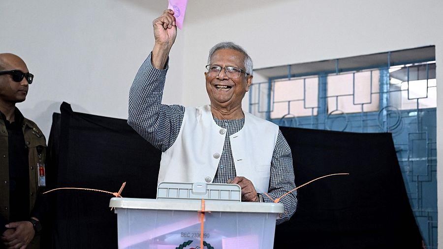 Un hombre sonriente deposita una papeleta en una urna transparente. Viste camisa a cuadros y chaleco, y levanta la papeleta con la mano derecha. A su izquierda, otro hombre observa la escena.