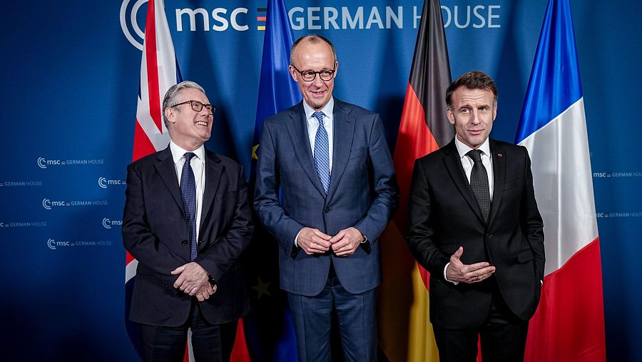 Tres hombres, vestidos con trajes y corbatas, posan frente a un fondo azul con un logotipo repetido. Cada uno está frente a una bandera diferente: Reino Unido, Unión Europea y Alemania, y Francia. La imagen fue tomada en un evento en Alemania.