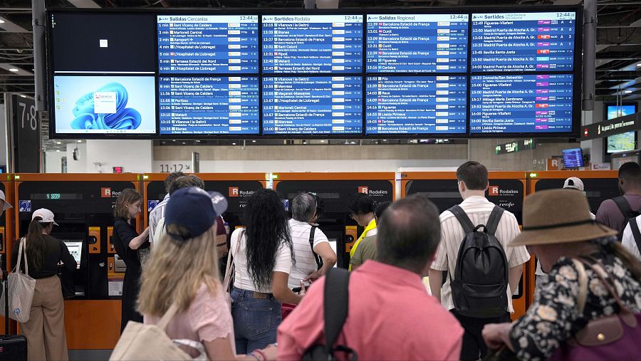 En una estación de tren, un grupo de personas espera frente a máquinas expendedoras. Carteles electrónicos exhiben información de salidas, incluyendo destinos como Sant Vicenç de Calders y Barcelona Estació de França.