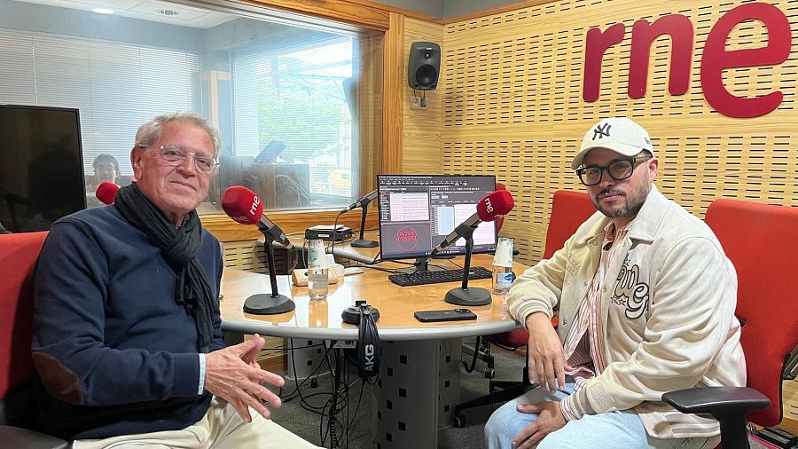 En un estudio de radio, dos hombres están sentados frente a una mesa con micrófonos y equipos de audio. Uno lleva una gorra y chaqueta con bordados, el otro un jersey oscuro y bufanda.