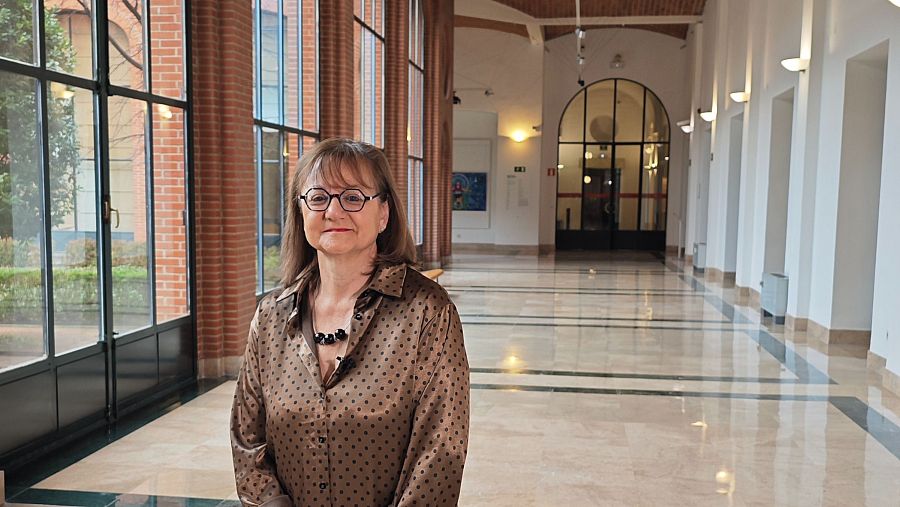 En la imagen, una mujer de mediana edad con gafas y cabello castaño claro, vestida con una blusa con lunares, se encuentra en el interior de un edificio, posiblemente un pasillo con suelo de mármol y ventanales.