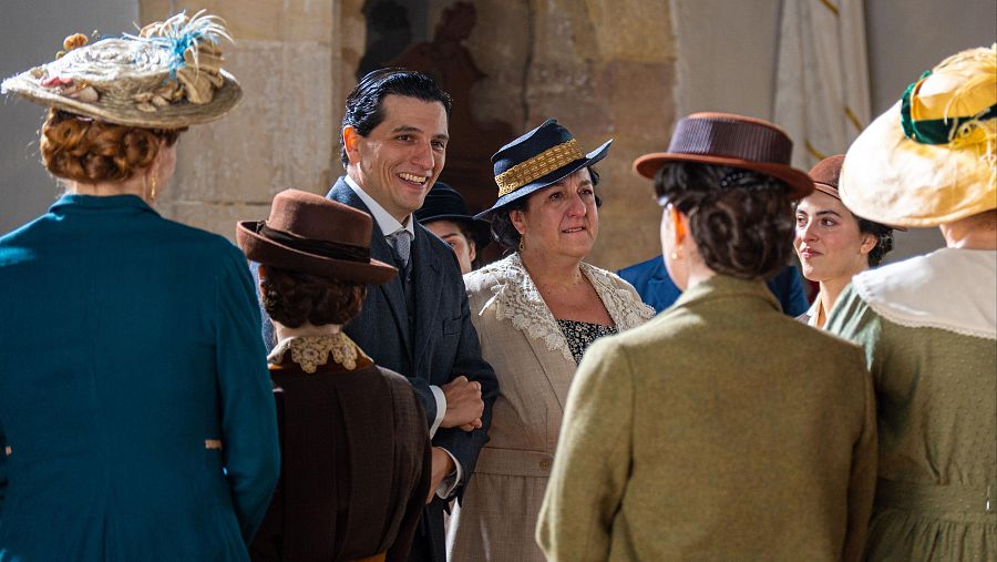 En una escena de época, un hombre sonriente con traje y corbata está junto a una mujer con vestido claro y sombrero. Otros personajes, vestidos con atuendos de la época, completan la escena en un interior.