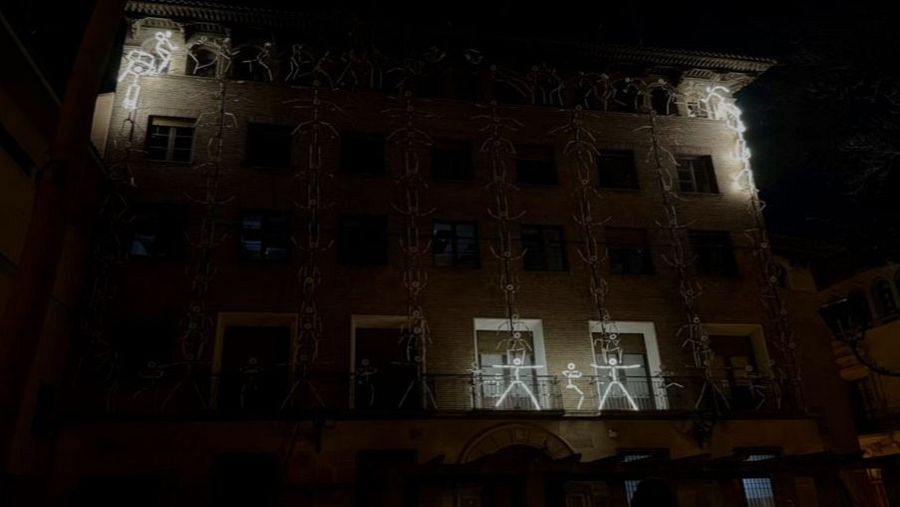 Fachada nocturna de un edificio de ladrillo con figuras luminosas esquemáticas que representan siluetas humanas, algunas de mayor tamaño en un balcón, creando un ambiente tenue.