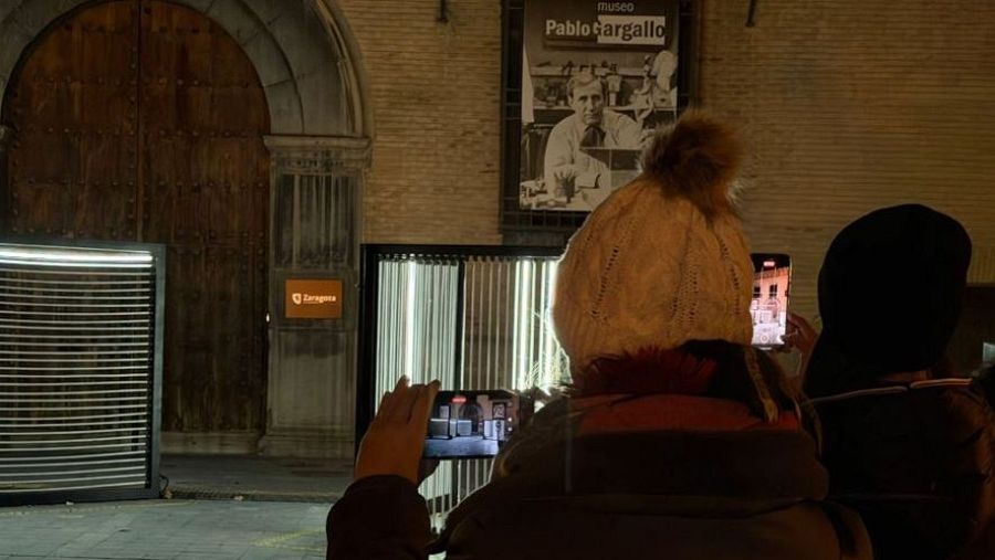 En la Plaza de San Felipe, dos individuos fotografían un edificio con sus móviles. Uno lleva un gorro crema y el otro una gorra negra, mientras que al fondo se distingue la fachada iluminada.