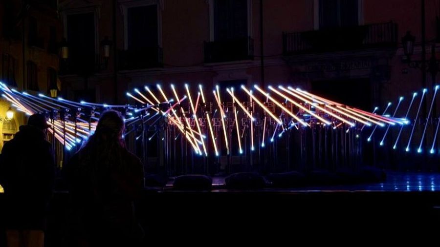 Dos figuras observan una instalación de luces abstractas en una plaza iluminada por la noche. Las luces, en tonos blancos y naranjas, contrastan con la arquitectura del entorno.