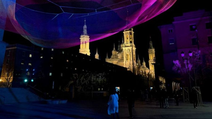 Vista nocturna de la Plaza de San Juan de los Panetes, con la silueta iluminada de una basílica al fondo y una estructura luminosa sobre la plaza. Hay personas caminando y disfrutando del ambiente festivo.