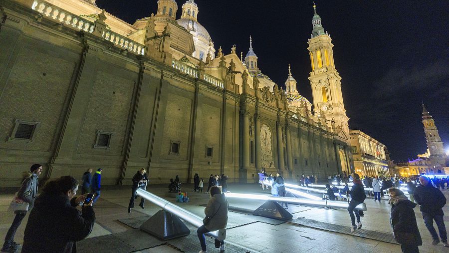 Una vista nocturna de la basílica, con sus cúpulas y torres iluminadas, domina la escena. En la plaza, una instalación de balancines con luz blanca atrae a personas que interactúan y toman fotos, sugiriendo un ambiente de ocio.