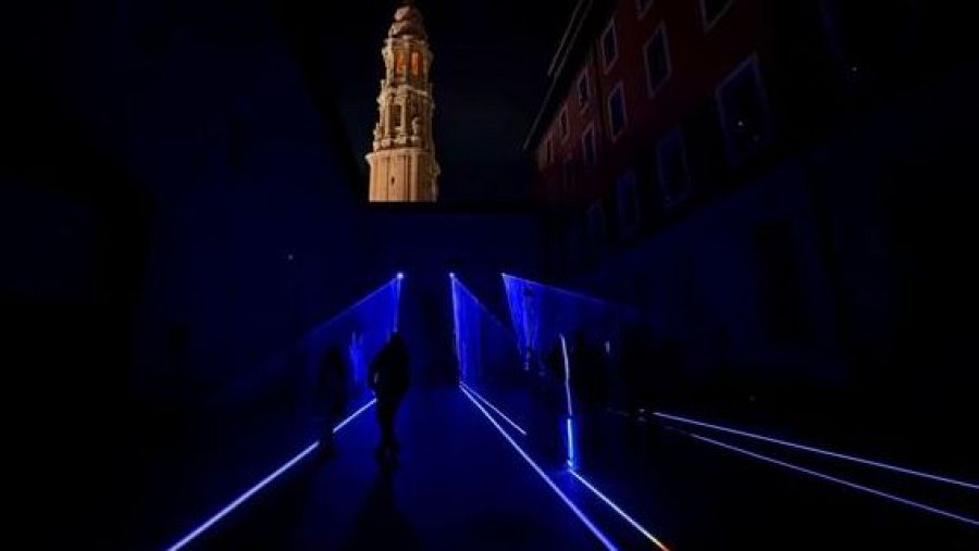 En el Patio diocesano del Museo, una instalación de luces azules guía a las siluetas de personas. La torre de un edificio religioso se alza en la oscuridad, creando una atmósfera misteriosa.
