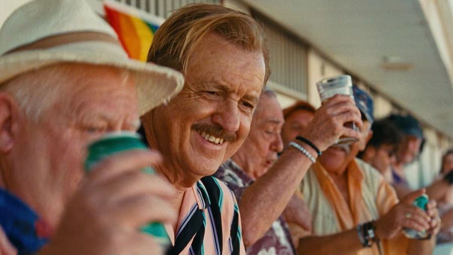 Un grupo de personas mayores celebra al aire libre, con un hombre sosteniendo una lata y otro sonriendo con una camisa a rayas. Se observan más personas con bebidas y una bandera ondeando.